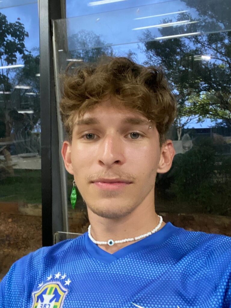 Sou um homem branco, de olhos verdes e cabelos loiros. Na foto estou de costas para uma janela, usando uma camiseta azul e um colar de pedras brancas.