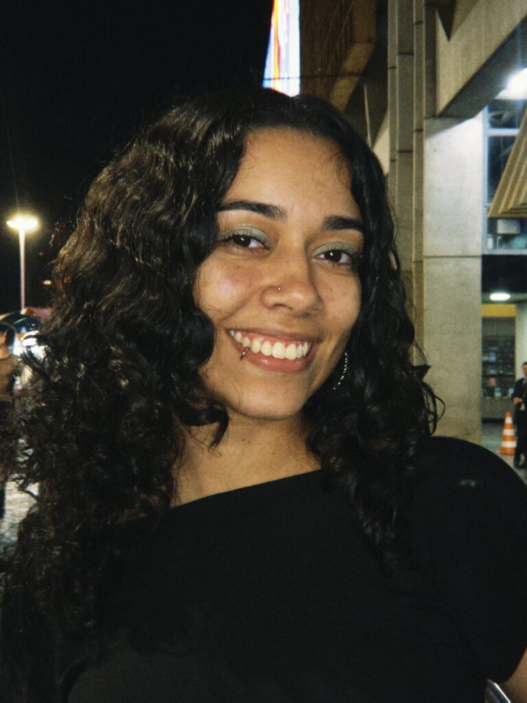 Sou uma mulher parda de cabelos cacheados e pretos, estou sorrindo para a câmera em um ambiente noturno. Na foto, uso uma blusa preta, tenho um piercing no lábio inferior e um pequeno piercing no nariz. Ao fundo, há pessoas caminhando e uma área urbana com luzes, incluindo letreiros e vitrines.