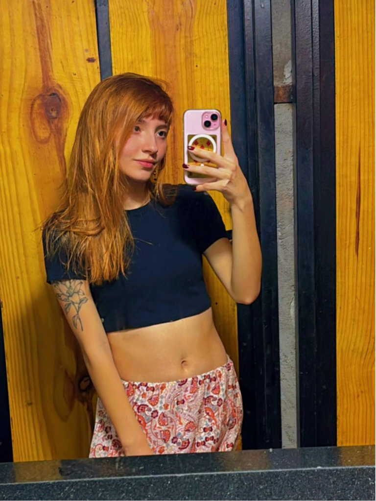 Sou uma mulher branca, olhos castanhos e cabelo ruivo. Na foto estou em frente a um espelho e olhando para o celular, usando uma blusa preta e uma saia florida. No meu braço, é possível ver uma tatuagem