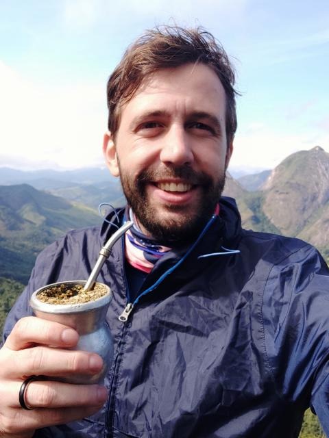 ​​Sou um homem branco, olhos cor verde e cabelo castanho. Na foto estou segurando uma cuia de chimarrão e ao fundo está uma cadeia de montanhas. Visto uma blusa azul e o meu cabelo está desarrumado.