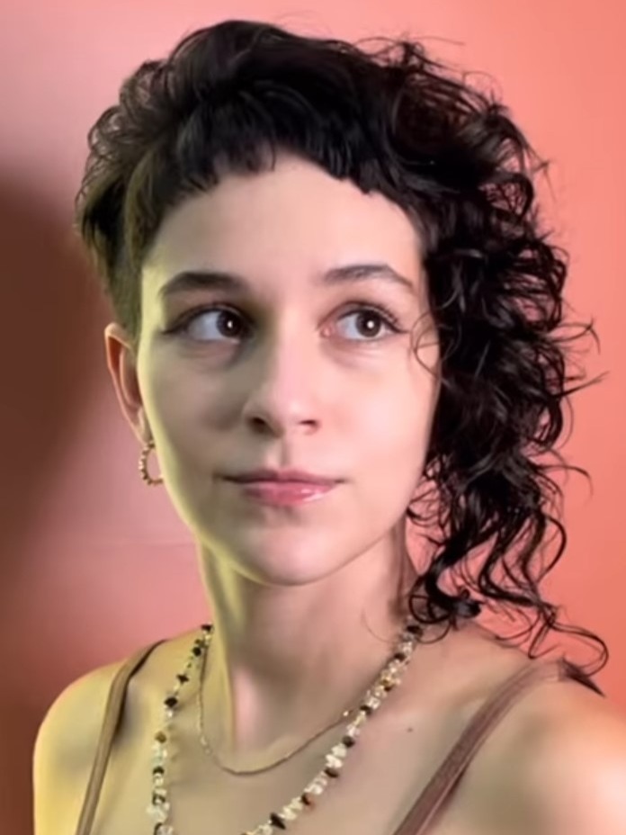 Sou uma mulher branca, na foto estou sob um fundo rosa e uso um colar de pedras verdes. Tenho olhos e cabelos castanhos e um corte de cabelo assimétrico.