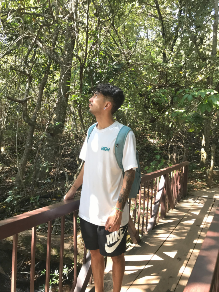 Sou um homem pardo vestido com uma blusa branca e short preto. Na foto estou em uma área verde posicionado de perfil, contemplando a natureza.
