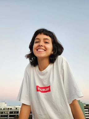 Sou uma mulher branca de cabelos pretos e lisos na altura dos ombros e com uma franja curtinha. Na foto estou usando uma camiseta branca larga com um retângulo vermelho no meio, sorrindo para a câmera e, atrás, dá pra ver o topo de alguns prédios e um céu claro iniciando o pôr do sol.