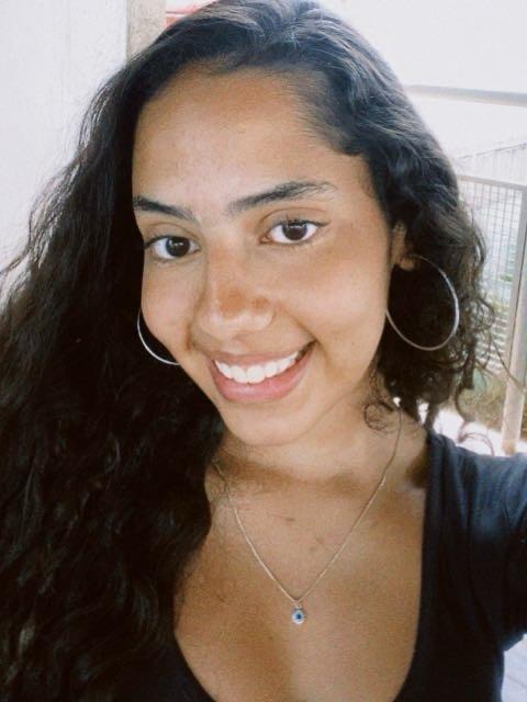 Sou uma mulher parda de cabelos cacheados e pretos, estou usando um brinco de argola e sorrindo para a foto. Na foto, uso uma blusa preta e um colar de olho grego.