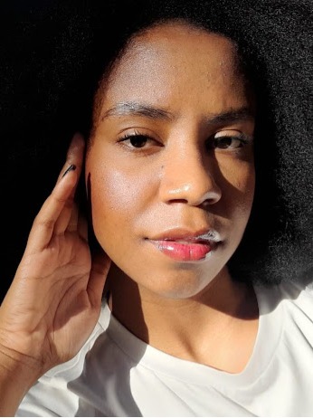 Sou uma mulher preta de cabelo crespo, na foto o ambiente está iluminado e estou olhando para a câmera com uma mão no rosto, usando uma camisa branca.