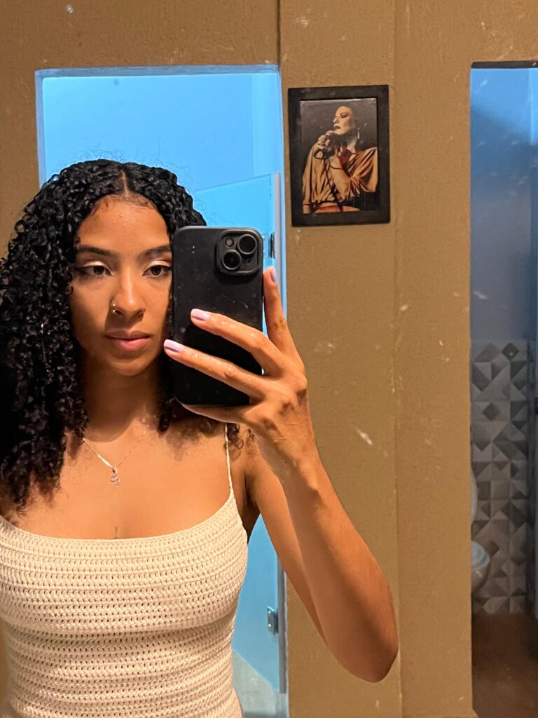 sou uma mulher preta de cabelo cacheado solto partido ao meio, vestida de branco, a foto foi tirada em frente ao espelho e estou com o celular com a capinha preta na mão e ao fundo é uma parede com um quadro de uma moça (não sei quem é)