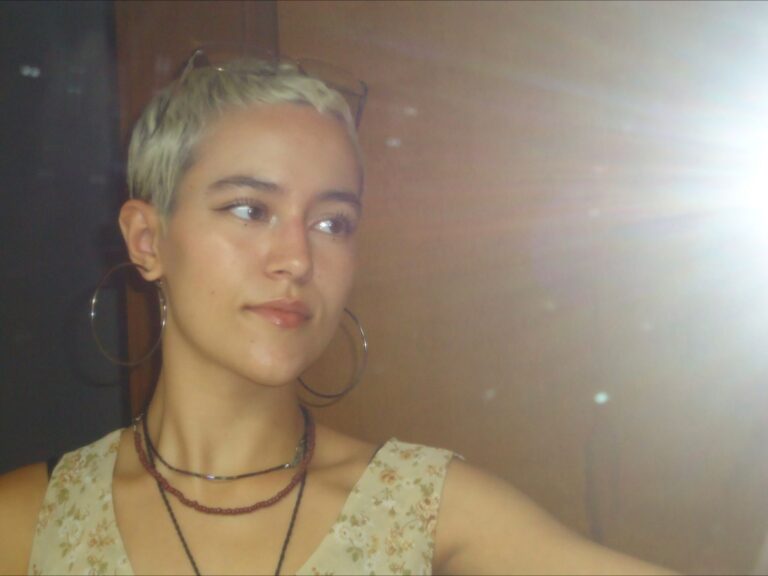 Sou uma mulher branca, tenho olhos castanhos e meu cabelo está bem curto, depois de ser raspado e descolorido. O flash da câmera se espalha pelo espelho onde a foto foi tirada. Estou usando brincos de argola grandes, 3 colares, uma regata clara e meus óculos de grau estão apoiados sobre a cabeça.